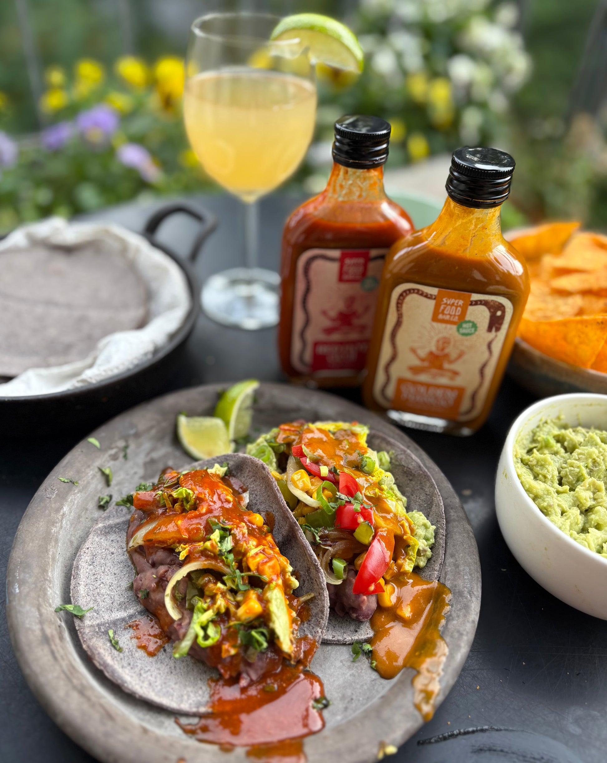 Table with a plate of tacos topped with Golden Mango and Red Hot Goji hot sauces, served with guacamole and tortilla chips